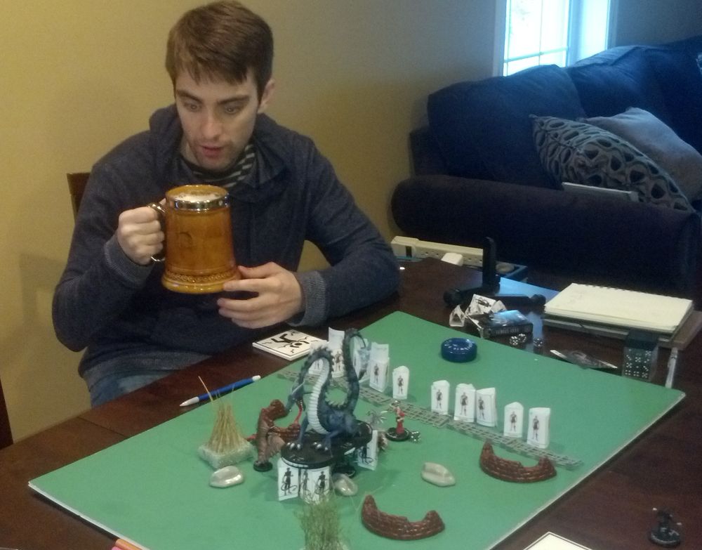Jeff at his dining table with a big stein, behind a green battle map with a dragon miniature and paper minis